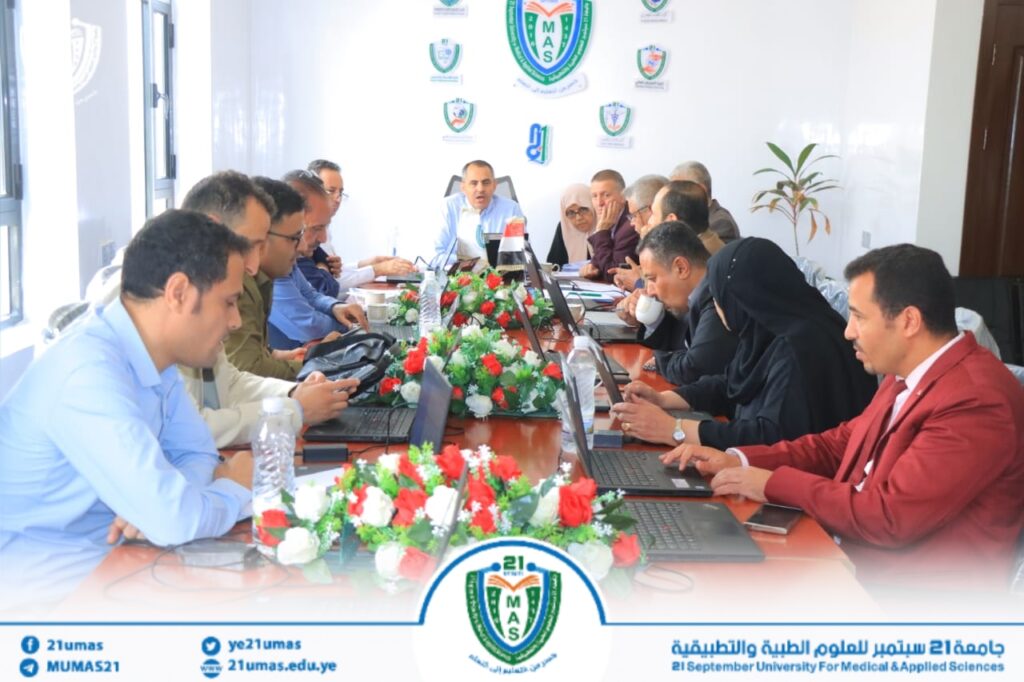Group of professionals around a long conference table with laptops, discussing in a meeting adorned with red‑white floral centerpieces and university logos on the wall.