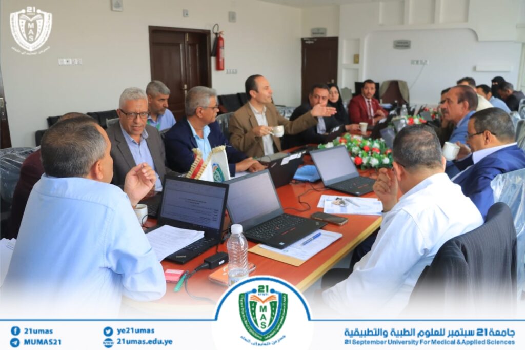 Group of professionals in a conference room around a long table with laptops, cups, and documents, featuring the university logo in the foreground artwork alternating with a floral centerpiece.
