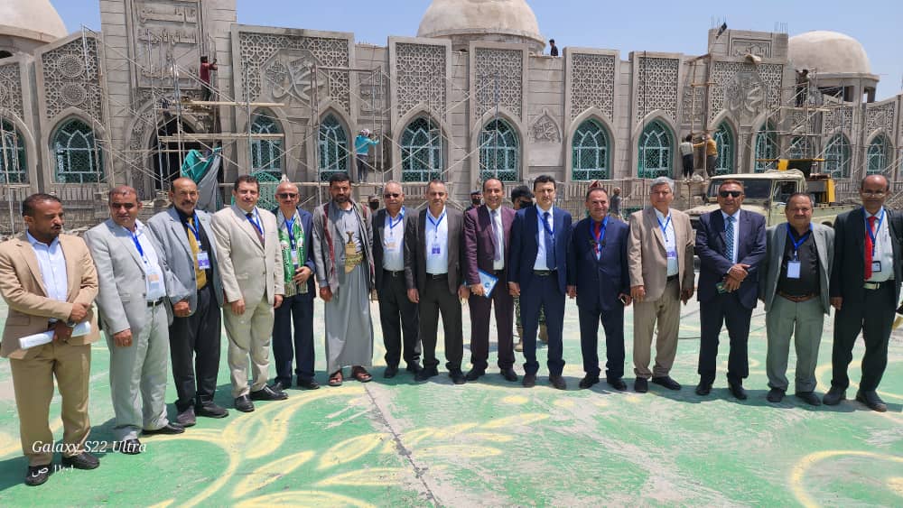 A group of men in suits posing for a photo in front of an ornate stone façade under construction, with scaffolding and workers in the background.