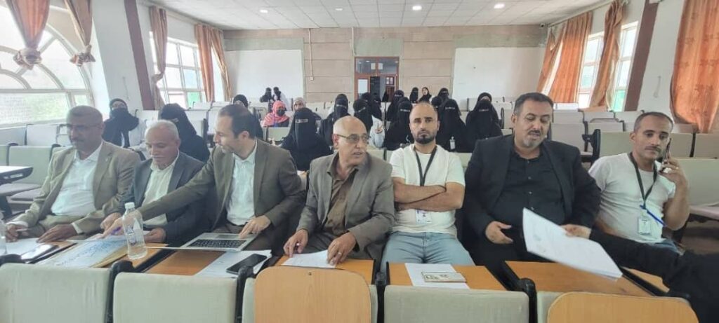 Front-row adults in a conference setting: men in suits with papers and a laptop, audience behind them in a classroom hall. Beside them, a water bottle sits on the table.