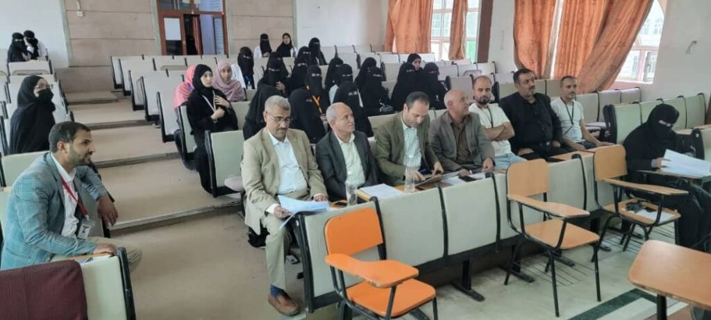 Front row of a lecture hall with several men in suits reviewing papers; women in hijabs seated in the background.