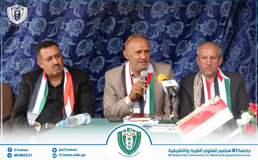 Three men in suits sit at a formal press conference table; the center man speaks into a microphone, wearing a scarf in tricolor (red, white, green) with two others beside him, a blue patterned backdrop behind them.