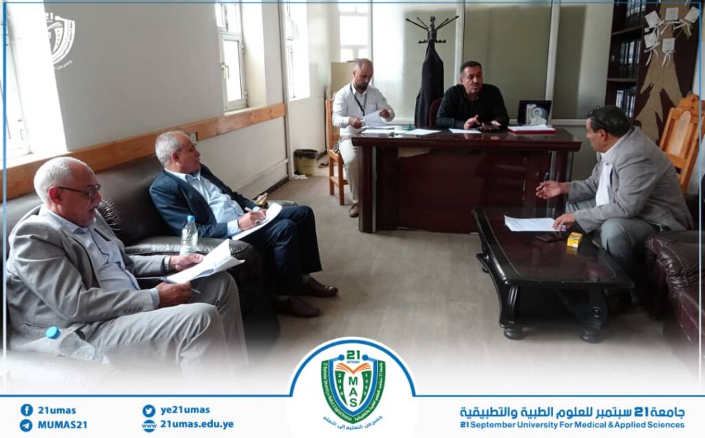 Five men in suits sit around a desk in a meeting room, papers and a water bottle on the table, a university crest visible on the banner at the bottom.