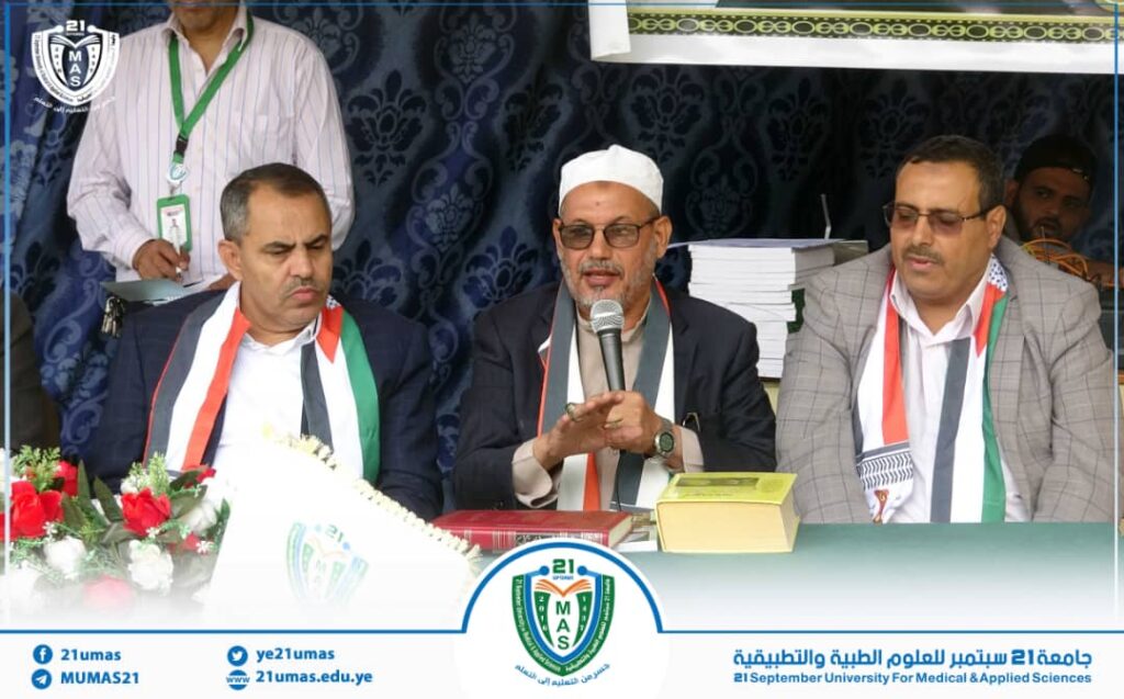 Three men sit at a panel table during a university event; the middle man speaks into a microphone, wearing a white cap and glasses with a scarf in national colors on his shoulders.