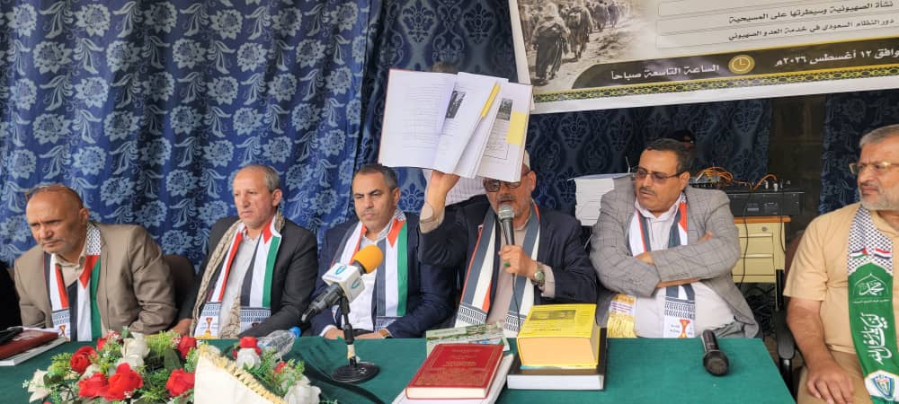 Six men sit at a press conference table with microphones, one raising a stack of papers while speaking; books, flowers, and scarves visible, blue patterned backdrop and Arabic banner behind them.