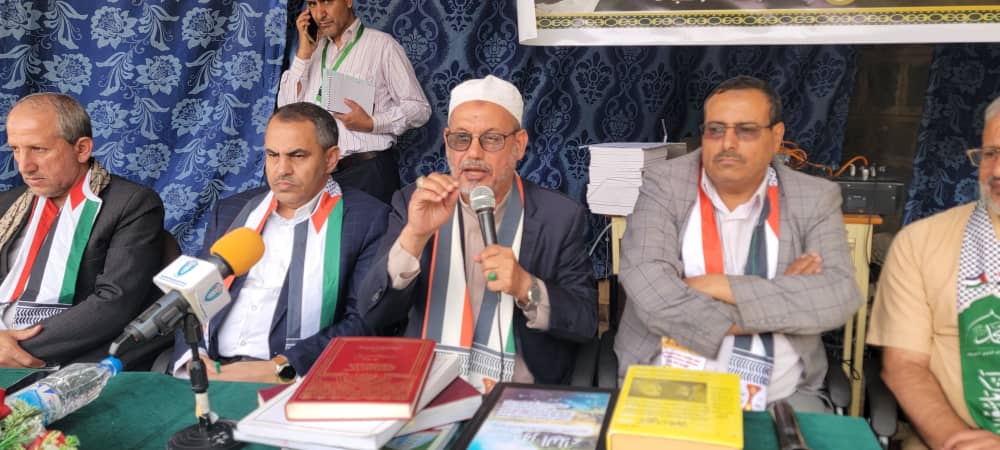 Group of men in suits seated at a table with microphones and books, one man speaking into a microphone at a panel. (Press conference)