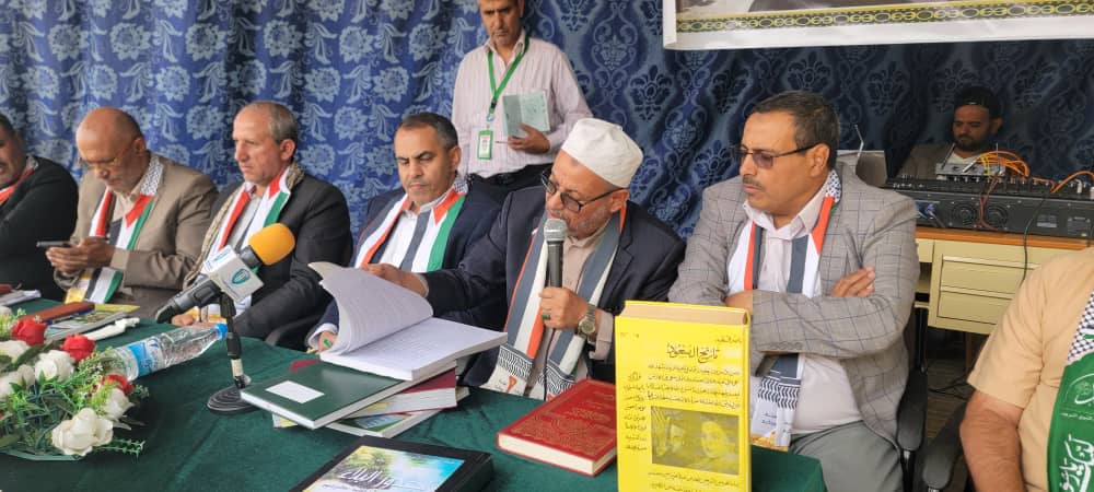 A group of men seated at a long table during a press conference, one man with a microphone reads from papers while others look on.
