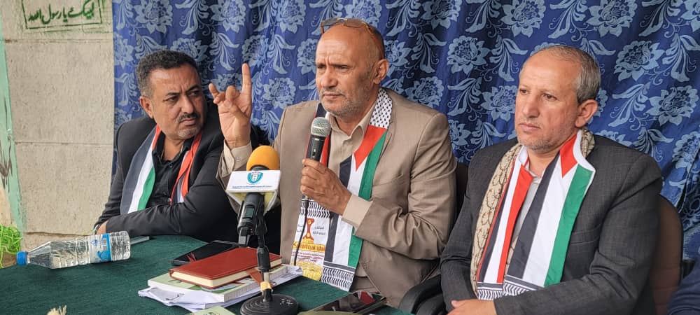 Three men in suits sit at a press conference table; the center man speaks into a microphone while the others listen, all wearing green-white-red scarves.