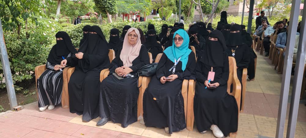 Group of women in black abayas seated outdoors at a gathering; two women wear turquoise and beige headscarves while others wear niqabs.