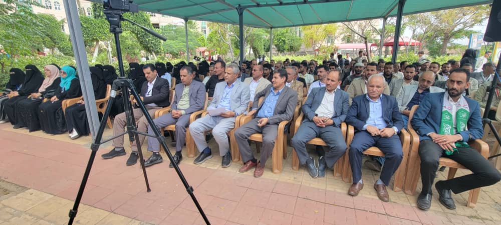 Audience seated under a teal canopy at an outdoor event; a camera on a tripod is in the foreground, people in suits and hijabs watching attentively.