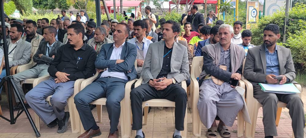 Group of men seated in plastic chairs at an outdoor event, listening attentively under a shaded area.