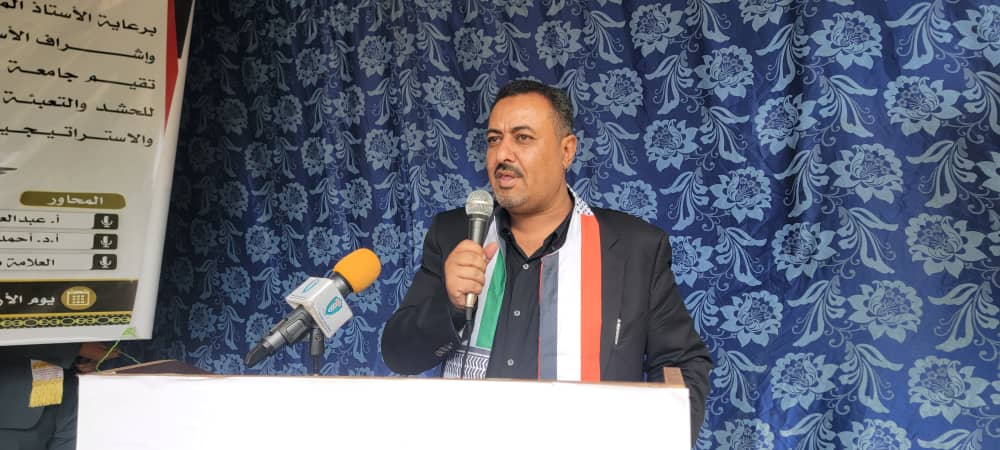 Man speaking at a podium with a microphone, wearing a tricolor sash against a blue floral backdrop.