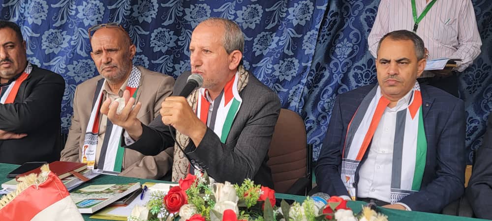 Man speaking into a microphone at a press conference table with two other men, all wearing scarves in Palestinian flag colors.