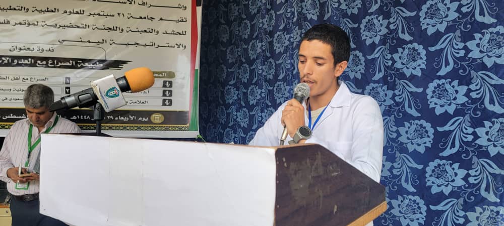 Young man in a white shirt speaks into a microphone at a podium, with a blue floral backdrop and a large Arabic banner behind him; another man stands to the left handling a device.