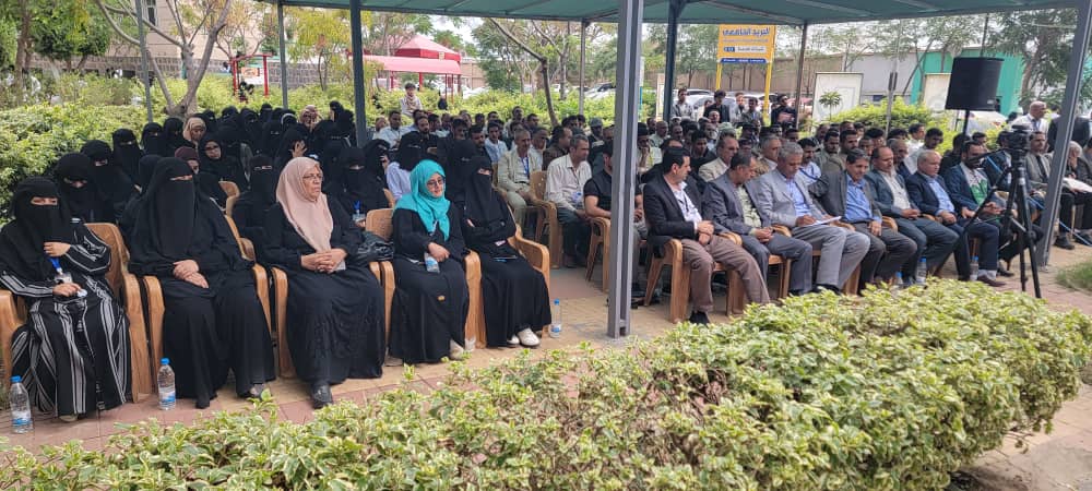 Outdoor event with rows of people seated under a canopy; many attendees in traditional attire, including women in abayas and niqabs, and men in suits. A camera tripod is visible to the right.