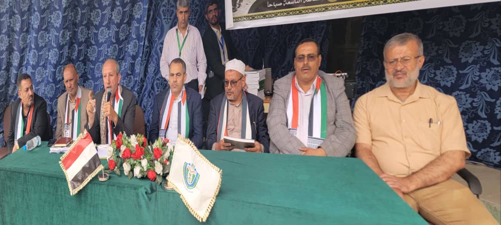 Panel of seven men seated at a green-cloth table with red-and-white flowers and flags, speaking at a formal event.