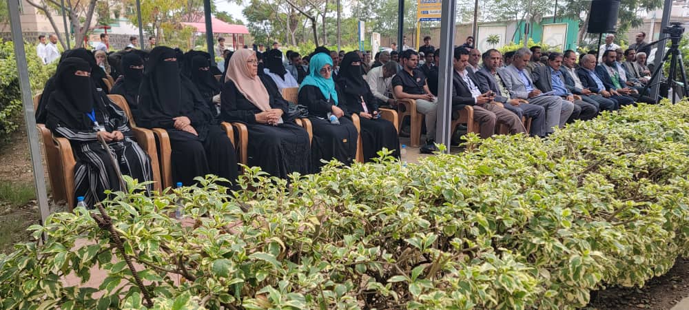 Audience seated outdoors at a ceremony; veiled women sit on the left and men on chairs to the right, with shrubs in front.