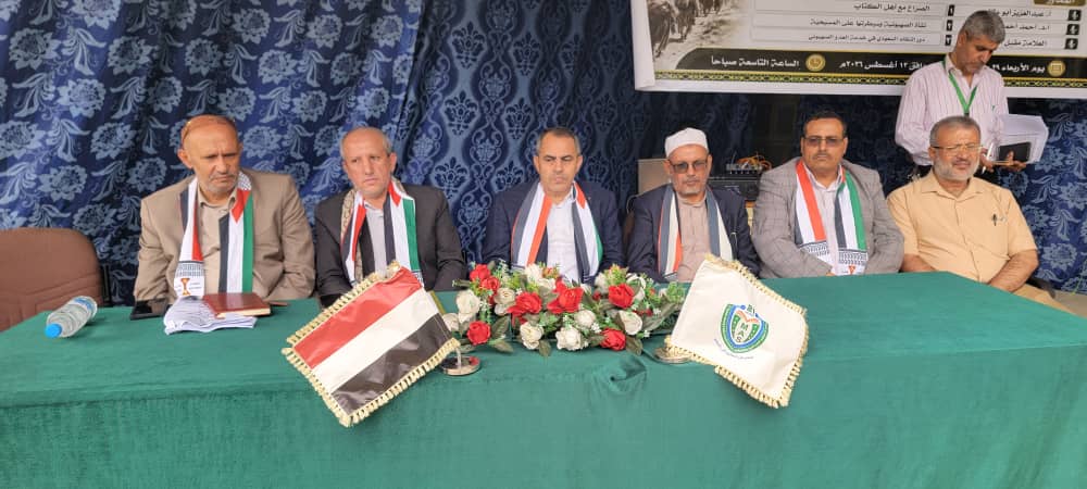 Six men seated behind a green conference table with a floral centerpiece and the Iraqi flag on display at a formal event.
