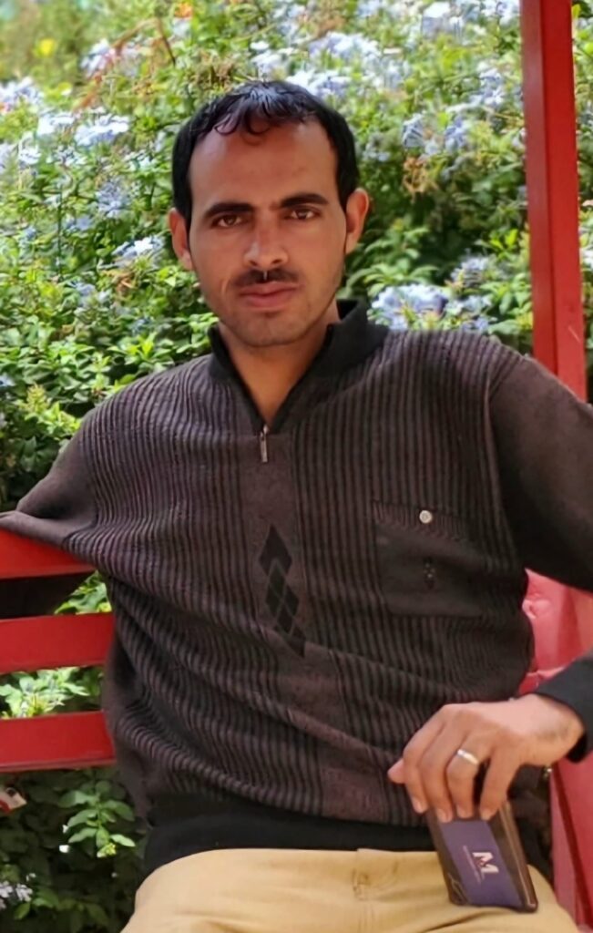 Man with dark hair and light facial hair sitting on a red bench outdoors, wearing a dark striped sweater, with greenery in the background.