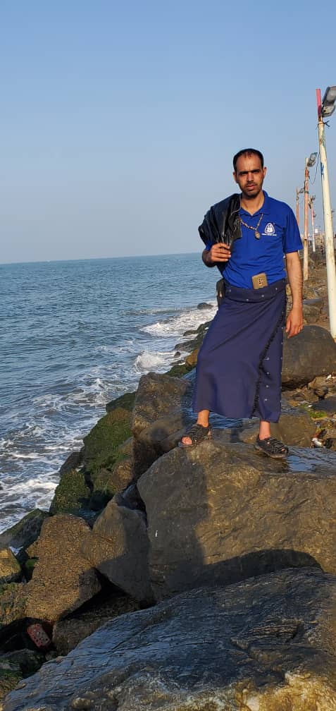 Man standing on rocky shoreline by the sea, wearing a blue polo and dark wrap, holding a jacket over his shoulder.