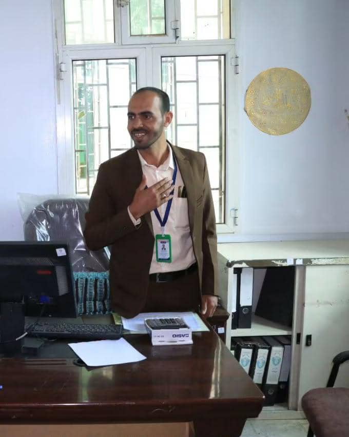 Man in a brown suit stands at a wooden desk in an office, hand over his chest, wearing an ID badge on a blue lanyard; a computer and file cabinet are in the background.
