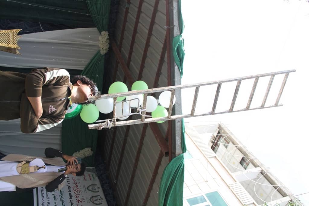 Man in a brown jacket speaks into a microphone at an outdoor event, with green/white balloons and a ladder on stage florida?