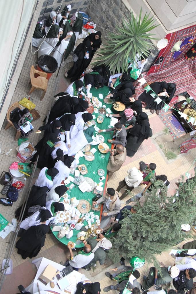 Top-down view of a large outdoor meal with women in black abayas and white head coverings sitting around a long green-cloth table sharing plates and bowls of food in a courtyard.