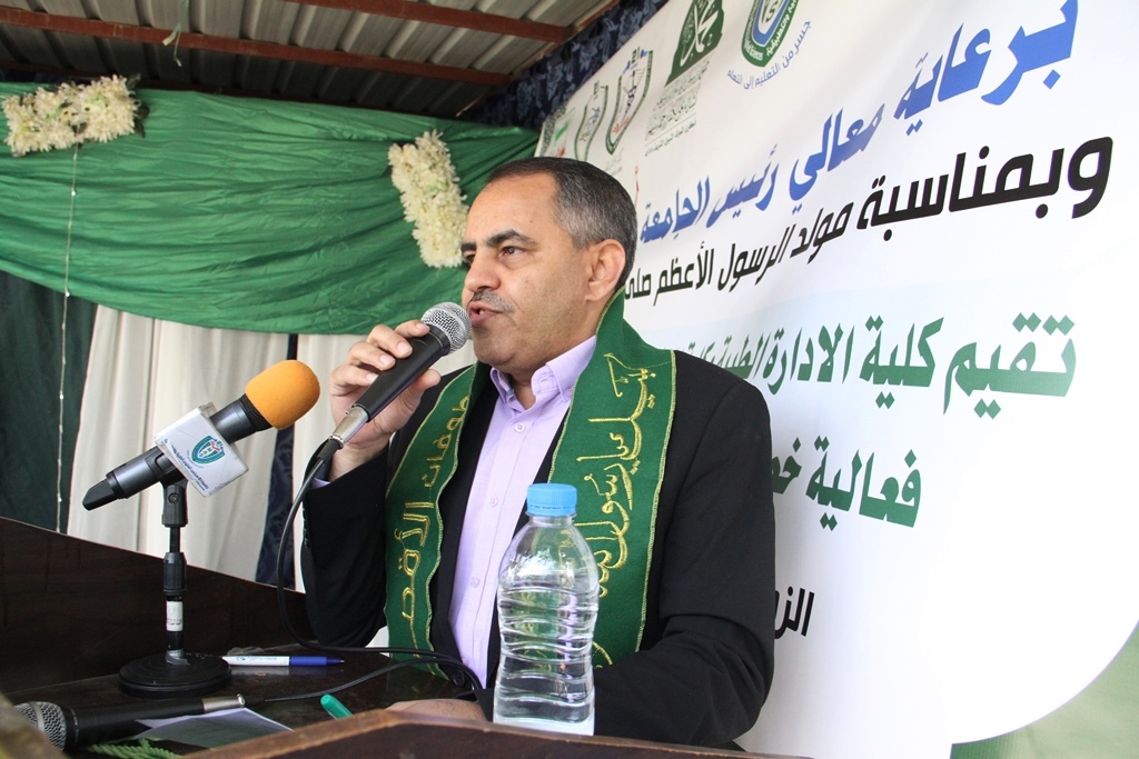 Man in a suit speaks into a microphone at a podium, wearing a green scarf, with Arabic banners in the background.