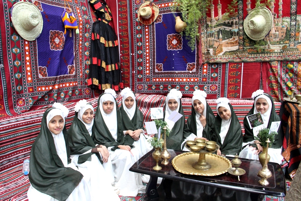 Group of young girls in white robes and green cloaks sit together in a colorfully decorated tent with rugs and hats around them, a brass tea set on the table nearby