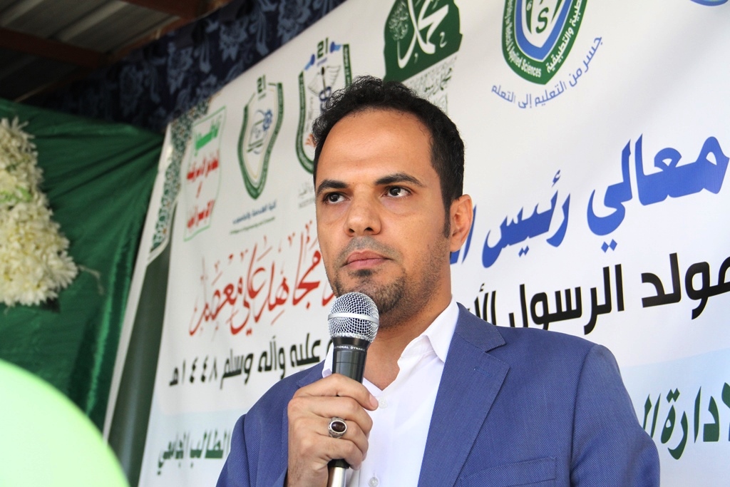 Man in a blue suit speaks into a microphone at an outdoor event with a large Arabic banner in the background.