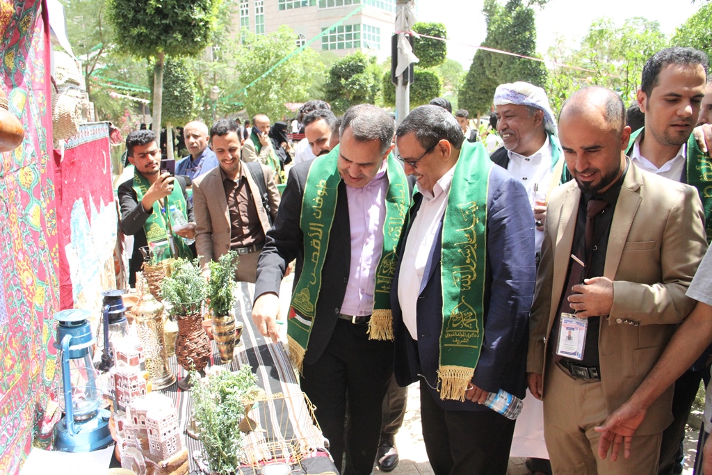 Group of men in suits with green sashes examining a display of potted plants, lanterns, and woven crafts at an outdoor festival.