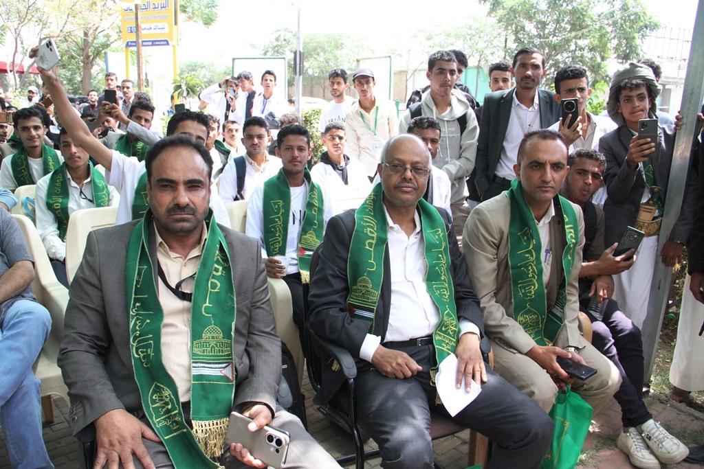 Group of men in suits with green sashes seated at an outdoor event, some taking photos with their phones.