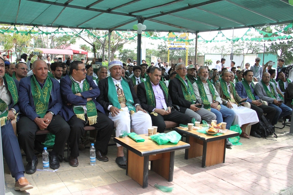 Group of men in suits wearing green ceremonial sashes seated in rows at an outdoor event under a shaded canopy, with snacks and drinks on tables nearby