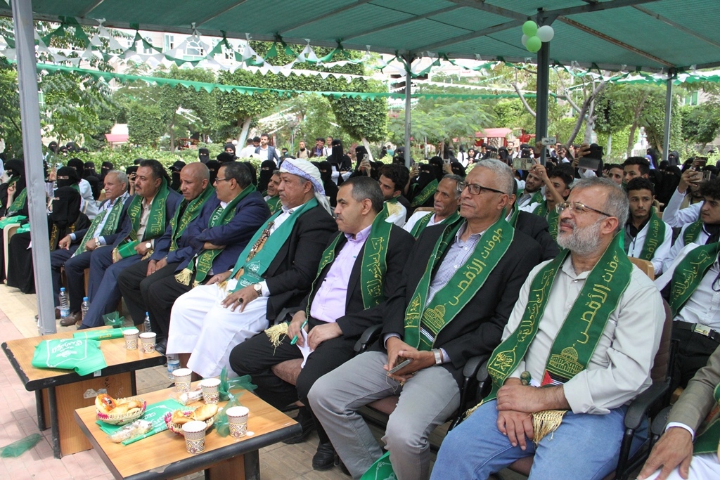 Group of formally dressed people seated under a green canopy at an outdoor ceremony, many wearing green ceremonial scarves with gold trim for a formal event.