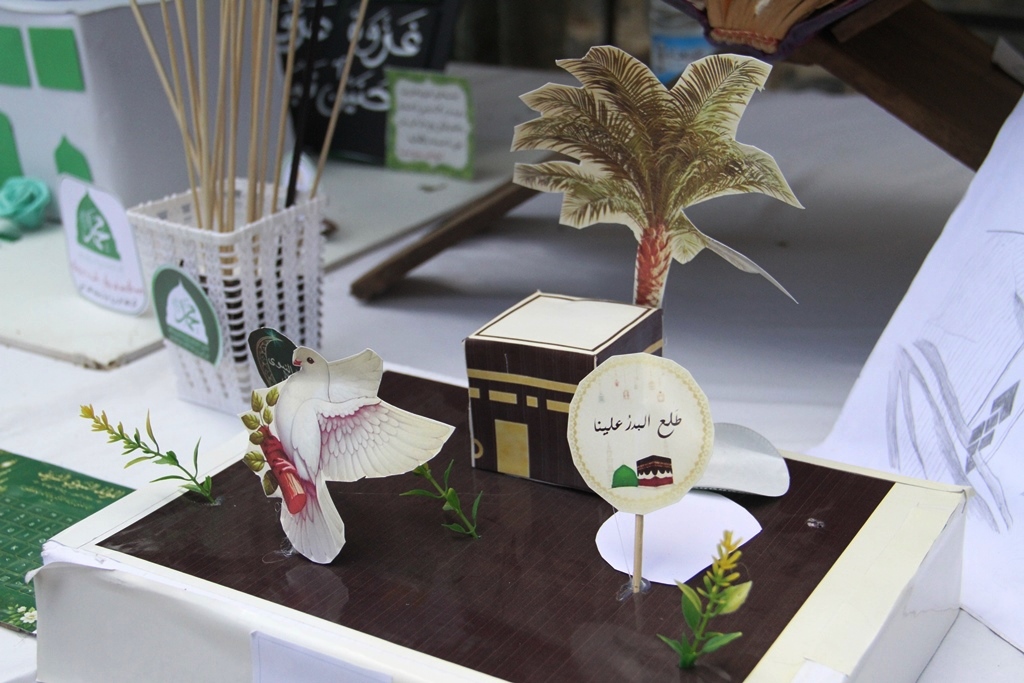 Paper craft diorama with a model Kaaba, a palm tree cutout, a white dove figure, and small plants on a dark base.