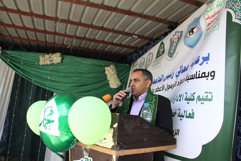 Man in a suit at a lectern giving a speech at an event, with green balloons and an Arabic banner in the background.