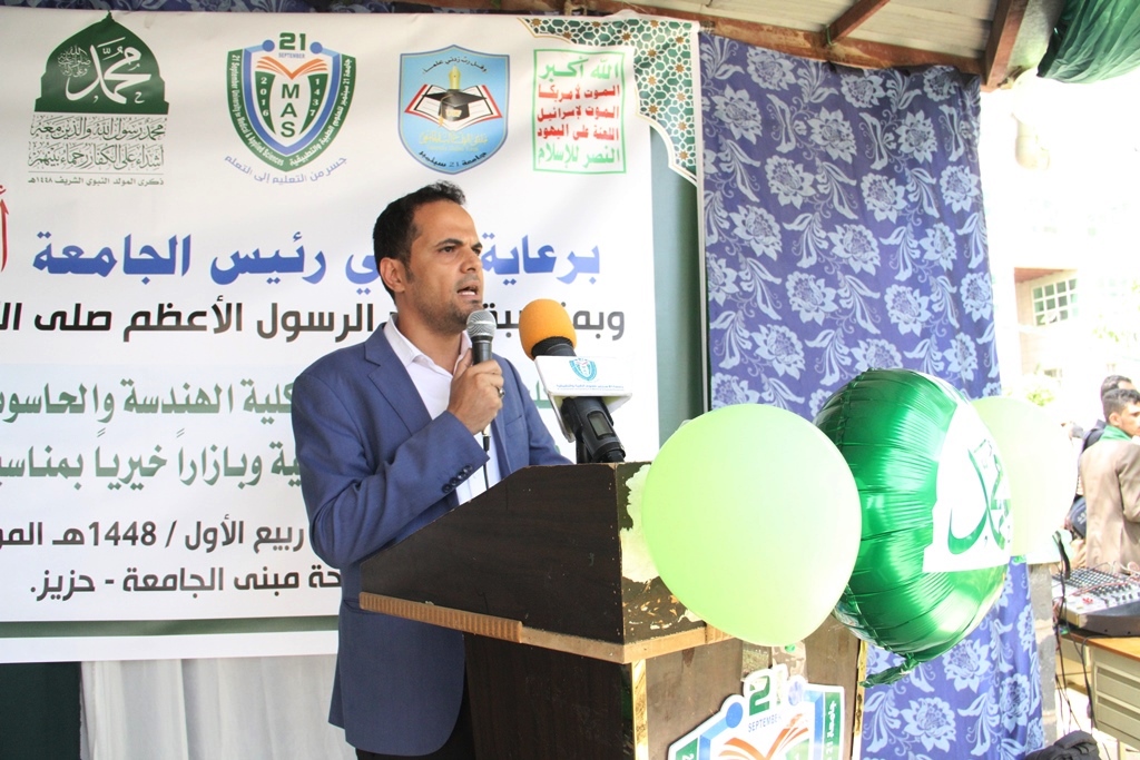Man in a blue suit speaks into a microphone at a podium during an Arabic university event, with banners and green balloons in the background.