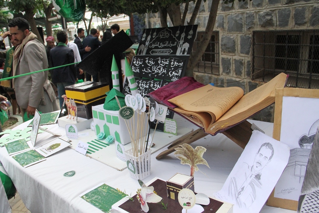 Outdoor stall with green-themed crafts and paper models on a white tablecloth, an open book on a stand, and people in the background.