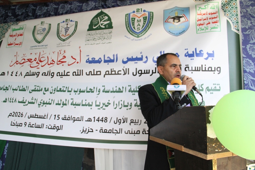Man in a suit speaks into a microphone at a podium during a ceremony, with a large Arabic banner and logos in the background.