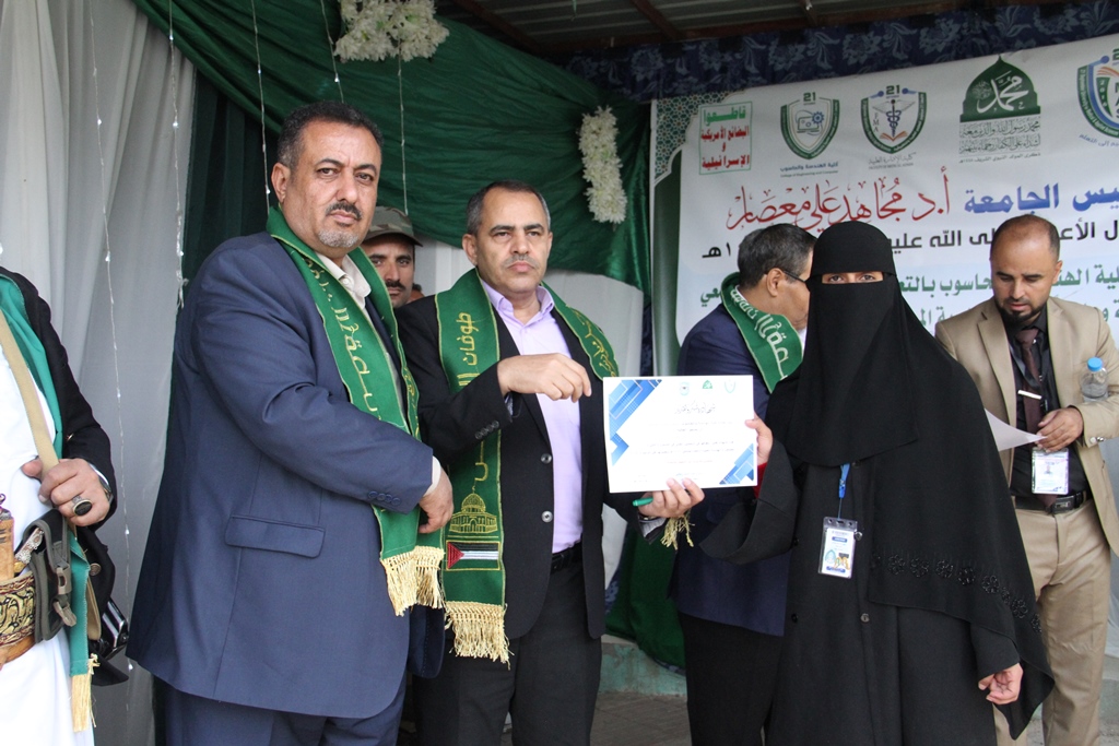 Officials wearing green scarves present a certificate to a woman in a black abaya at a ceremony, with an Arabic banner in the background.