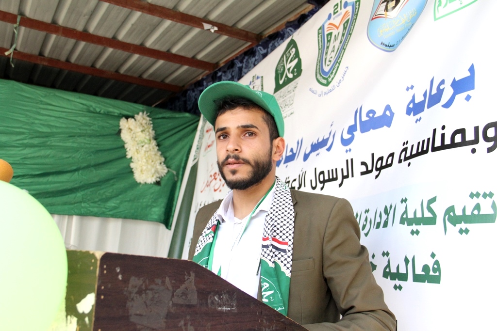 Young man in a green cap and brown blazer speaks at a podium during an event, with Arabic banners behind him.