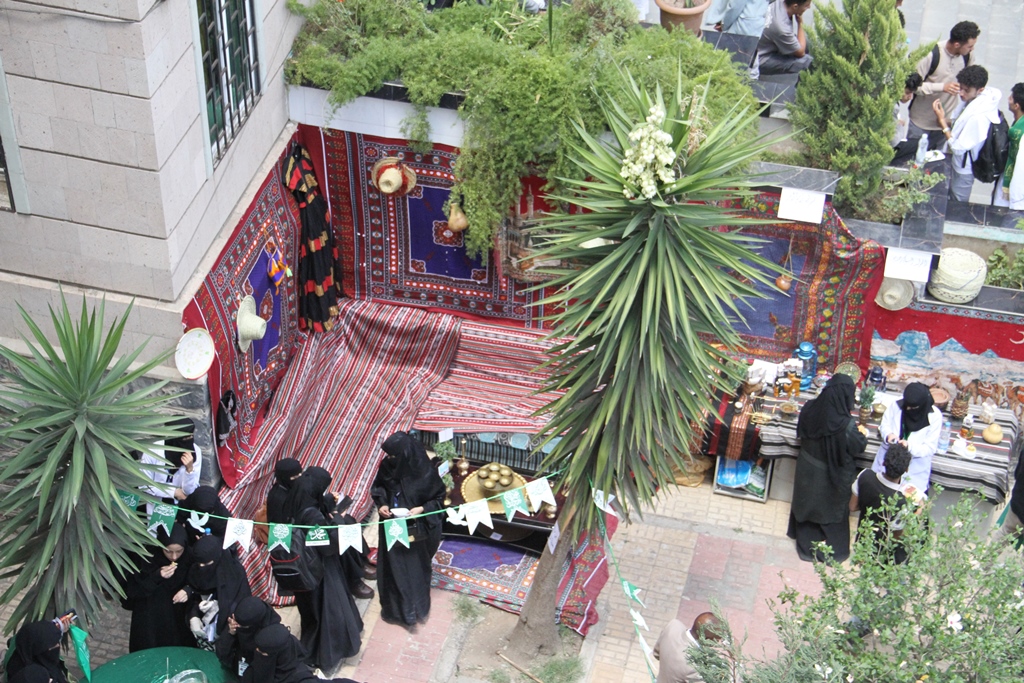 Outdoor market stall with red patterned carpets and textiles on display; women in black abayas browse wares under palm trees.