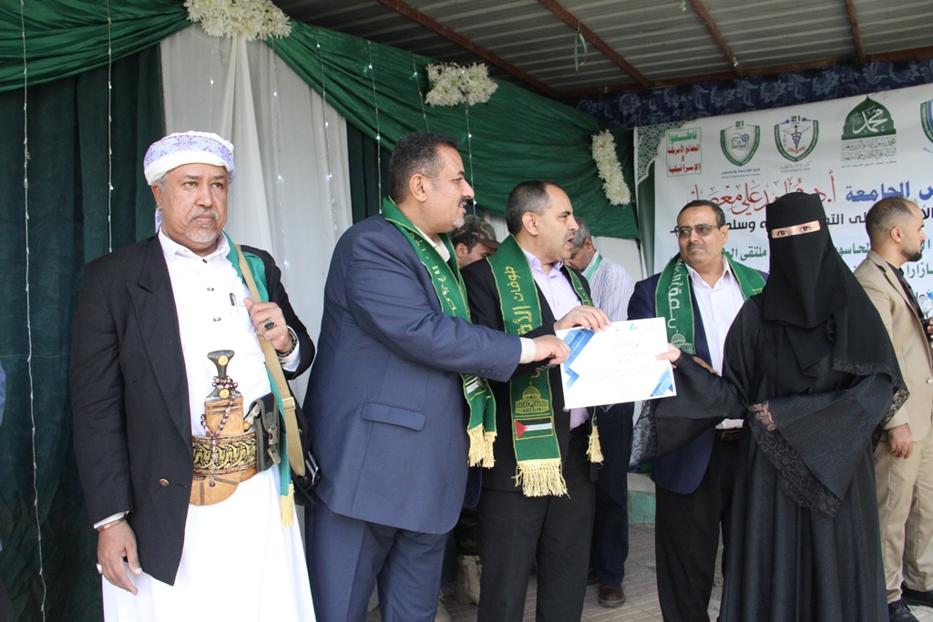 Group of men in suits and traditional dress award a certificate to a hijab-clad woman during a formal ceremony, with Arabic banners in the background.