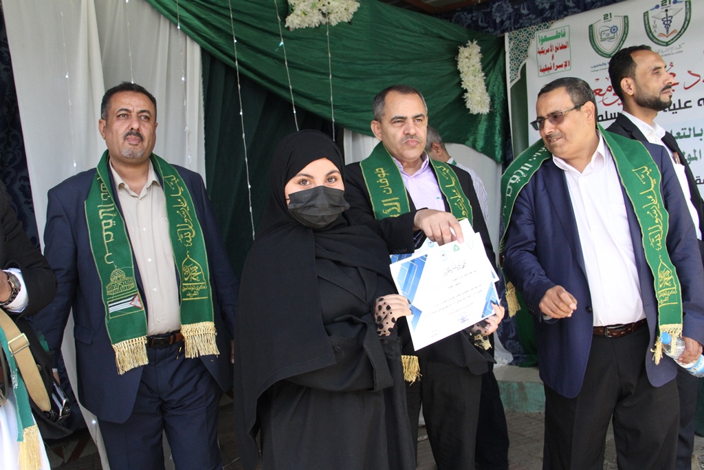 Group of formally dressed people on a stage, a woman in a black hijab and mask holds a certificate while others wear green sashes and smiles nearby.