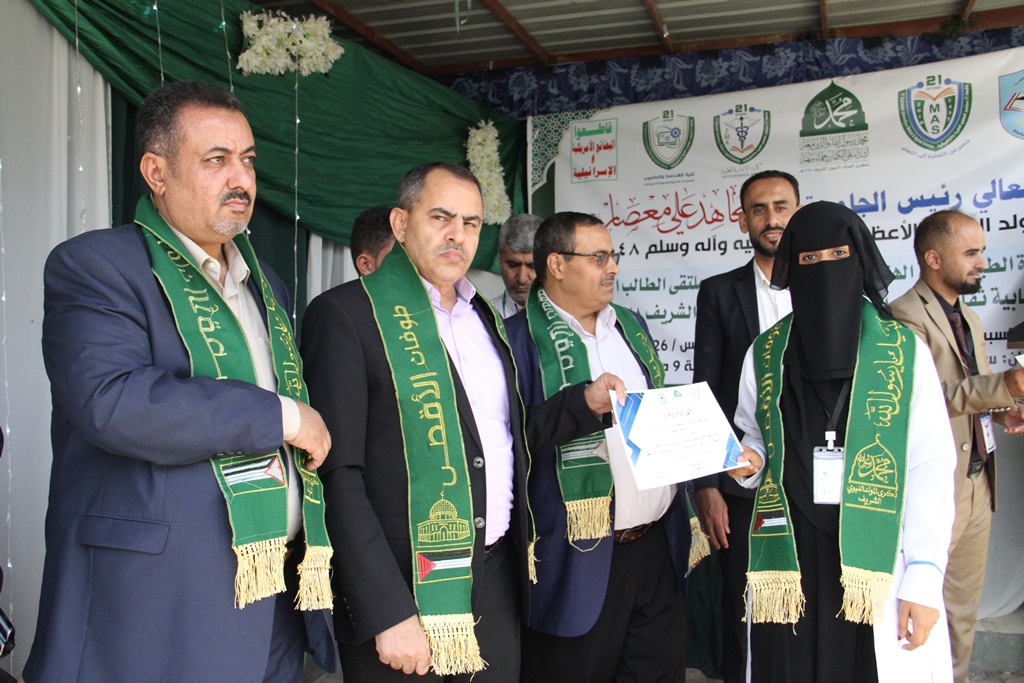 Group of men and a veiled woman on stage during an award ceremony, green sashes draped over their shoulders as a certificate is handed over in front of a banner.