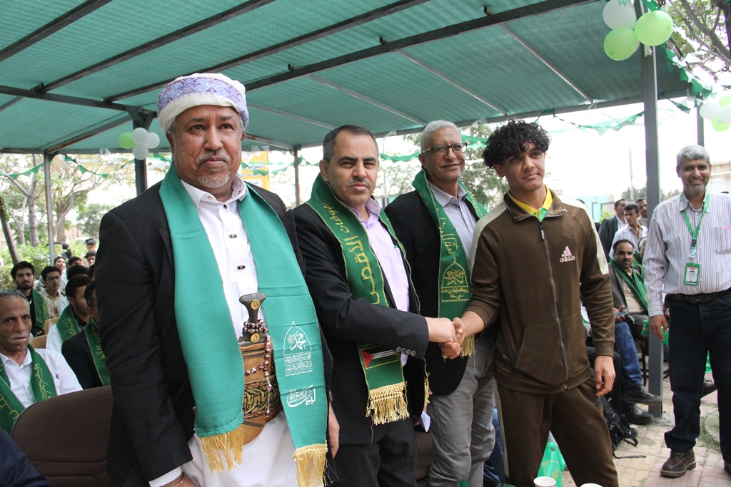 Group of men at an outdoor ceremony; a man in traditional attire shakes hands with a young man as others look on wearing green sashes and lanyards.