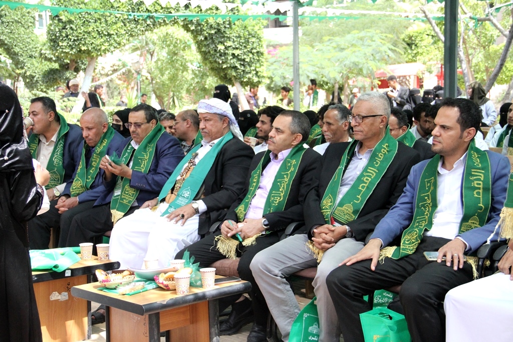 Group of men in suits with green ceremonial sashes seated outdoors at a formal event, a woman speaks at left.