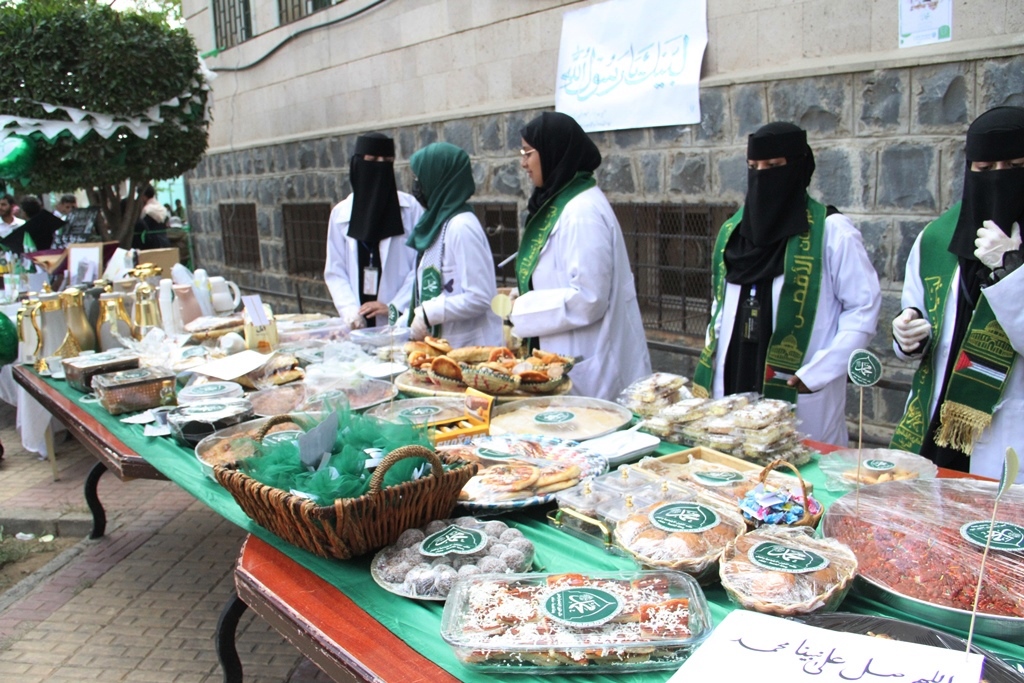 Women in white robes and green scarves stand behind a long table of assorted baked goods and snacks at an outdoor stall.