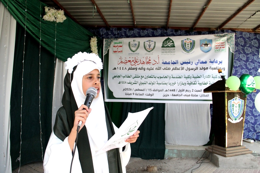 Young girl in a white hijab and black cloak speaks into a microphone on a stage with a banner behind her at a formal event.