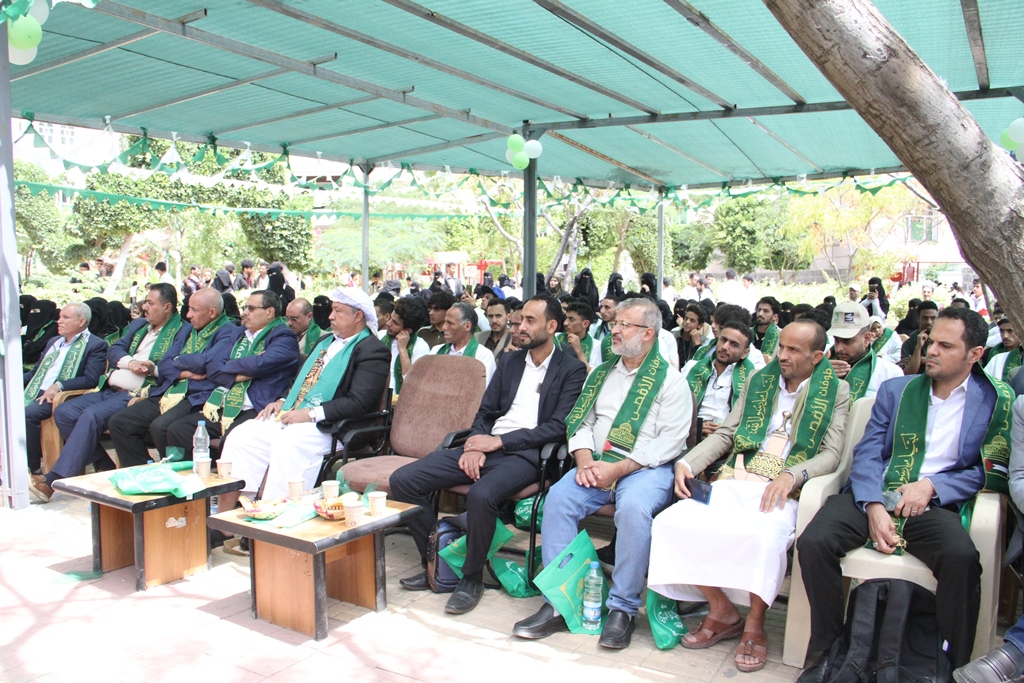 Outdoor ceremony with a large crowd seated under a green canopy; many attendees wear green sashes and formal attire.